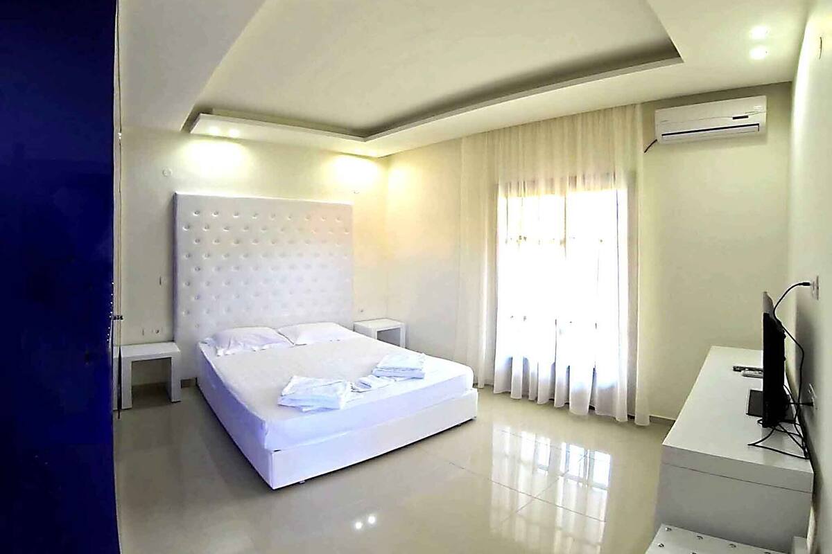 A modern hotel room with a large bed, a bedside table, an air conditioning unit, and a desk with a chair.