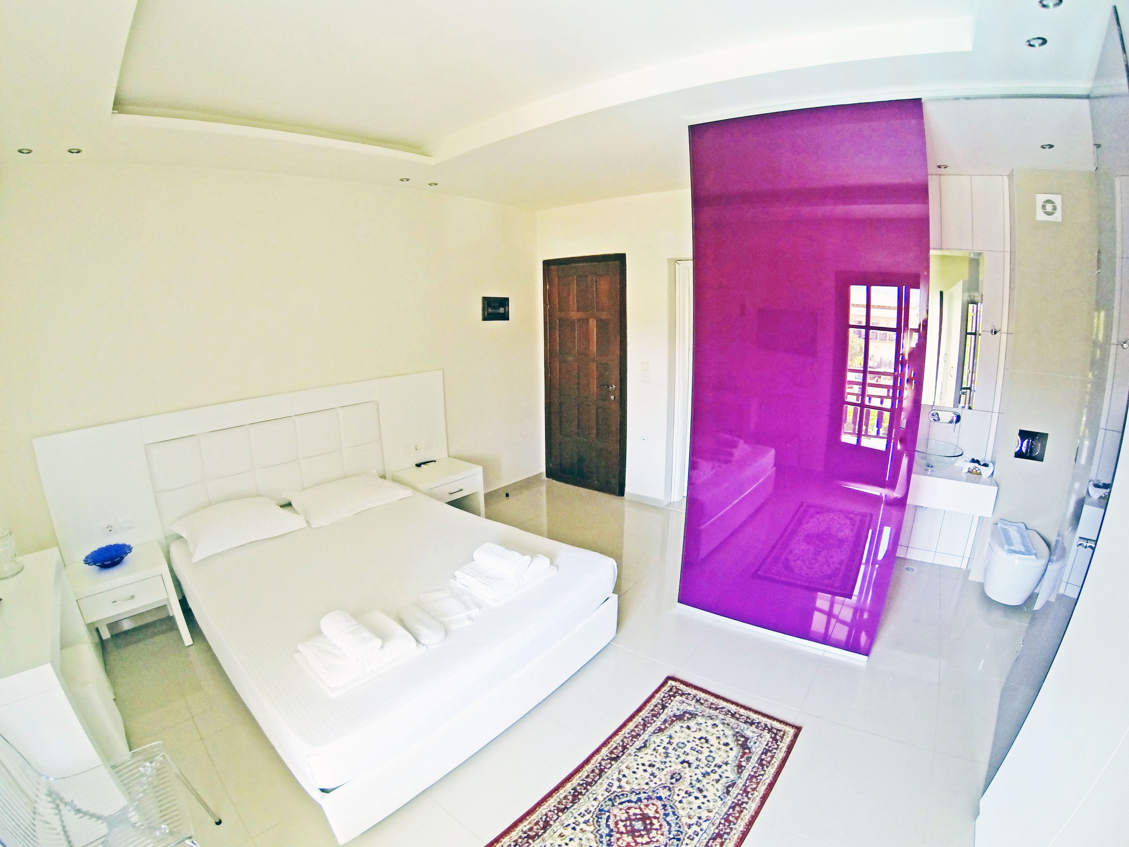 A modern hotel room with a large bed, a bedside table, a small desk, and a bathroom visible through a glass partition.