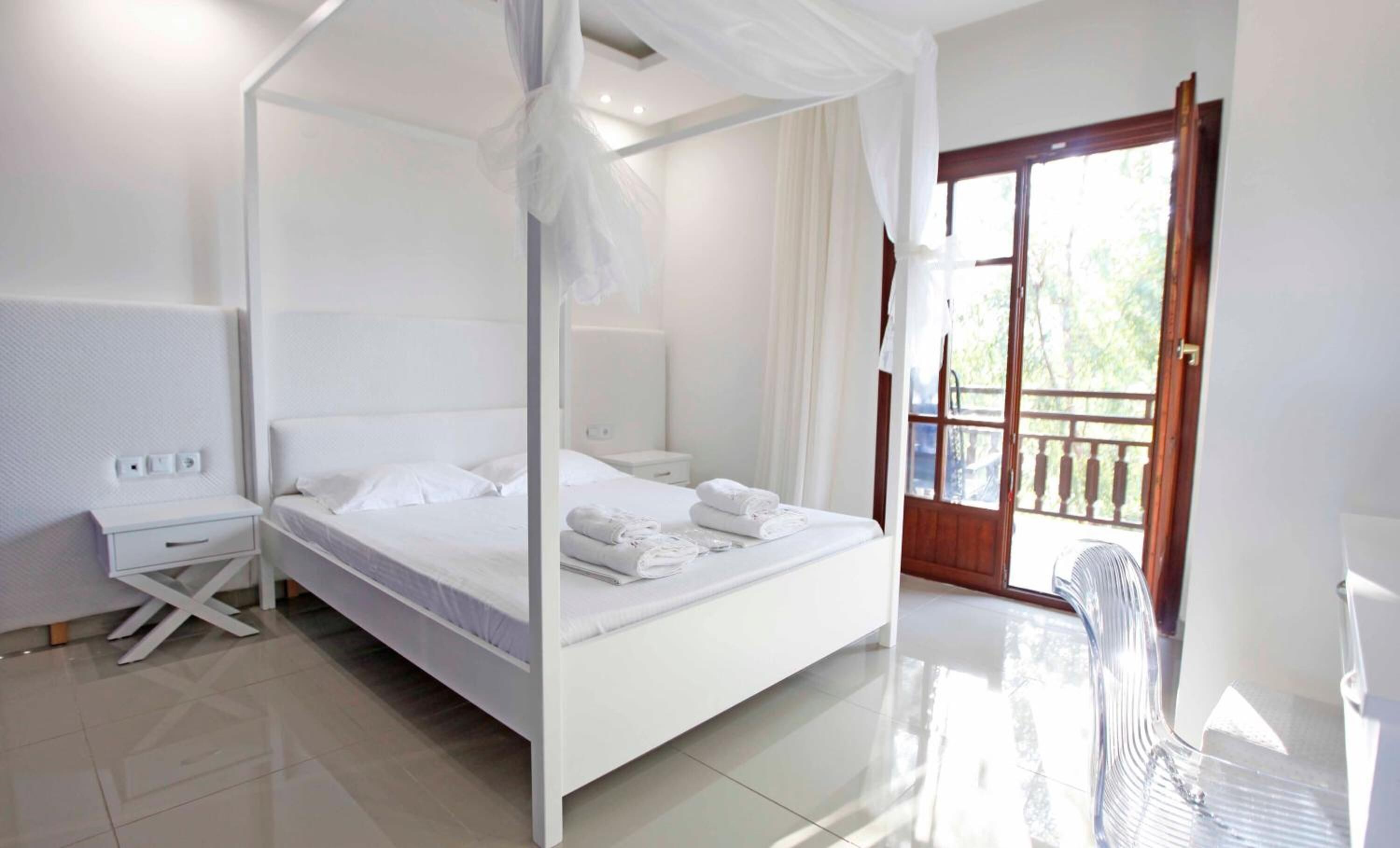 A four-poster canopy bed with white linens and towels, a bedside table, and a view of the outdoors through a glass door.