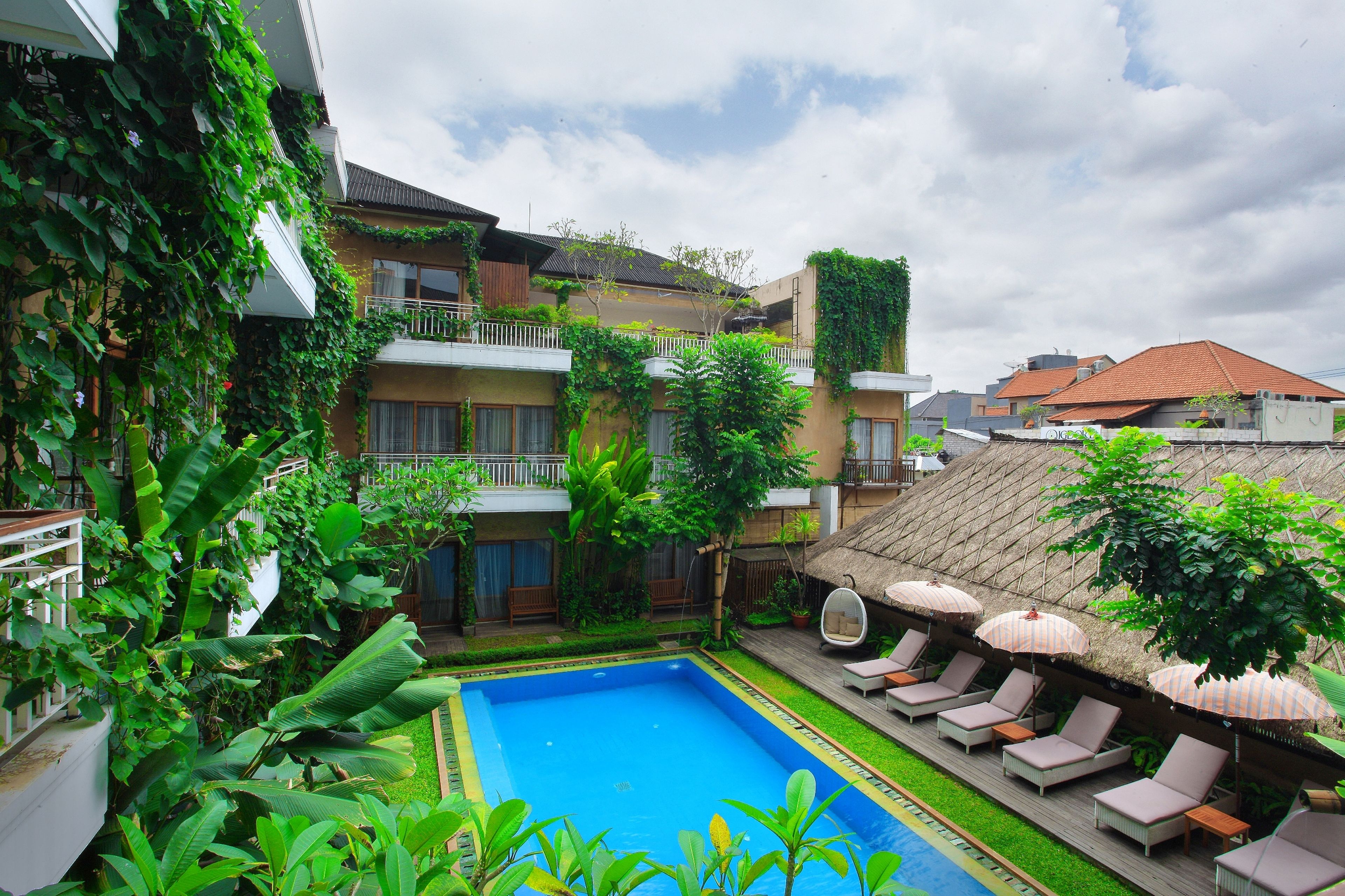 A swimming pool surrounded by lush greenery, a thatched-roof structure, and lounge chairs with umbrellas.