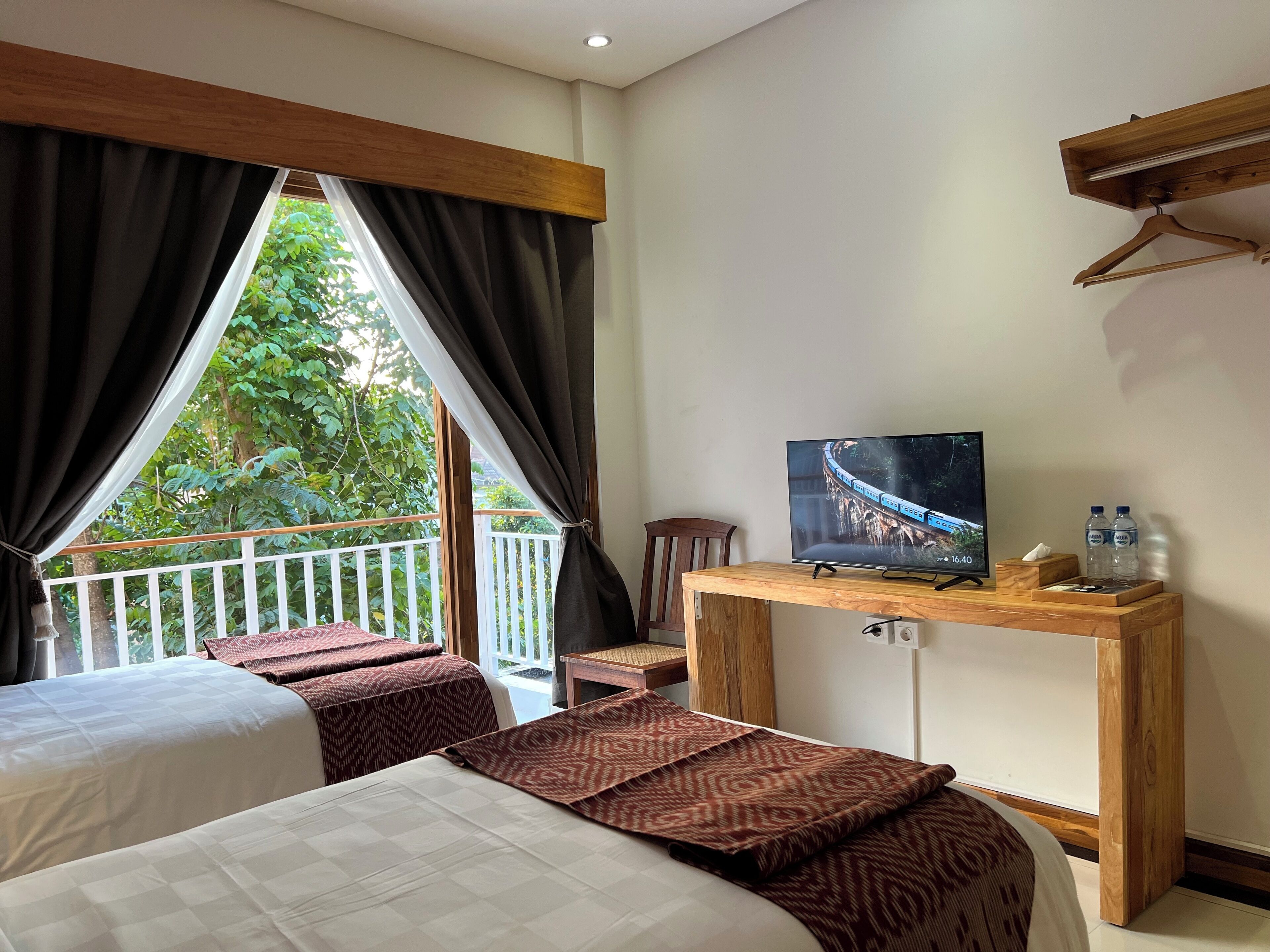 A hotel room with a bed, a small table with a TV, and a balcony with a view of greenery.