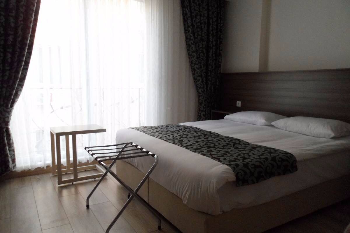 Una cama bien hecha con un edredón estampado, una mesita y un tendedero en una habitación con un ventanal y cortinas.