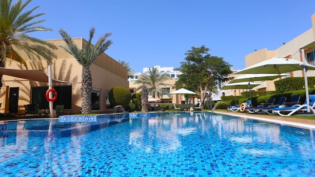 Una piscina con sillones de descanso y sombrillas, palmeras y un edificio al fondo.