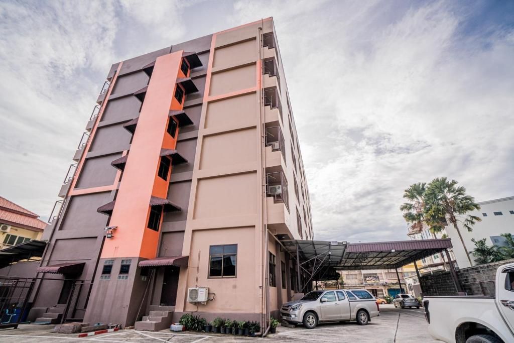 Un edificio de varios pisos con un elemento vertical naranja en el centro, una zona de estacionamiento cubierta y un vehículo blanco estacionado al frente.