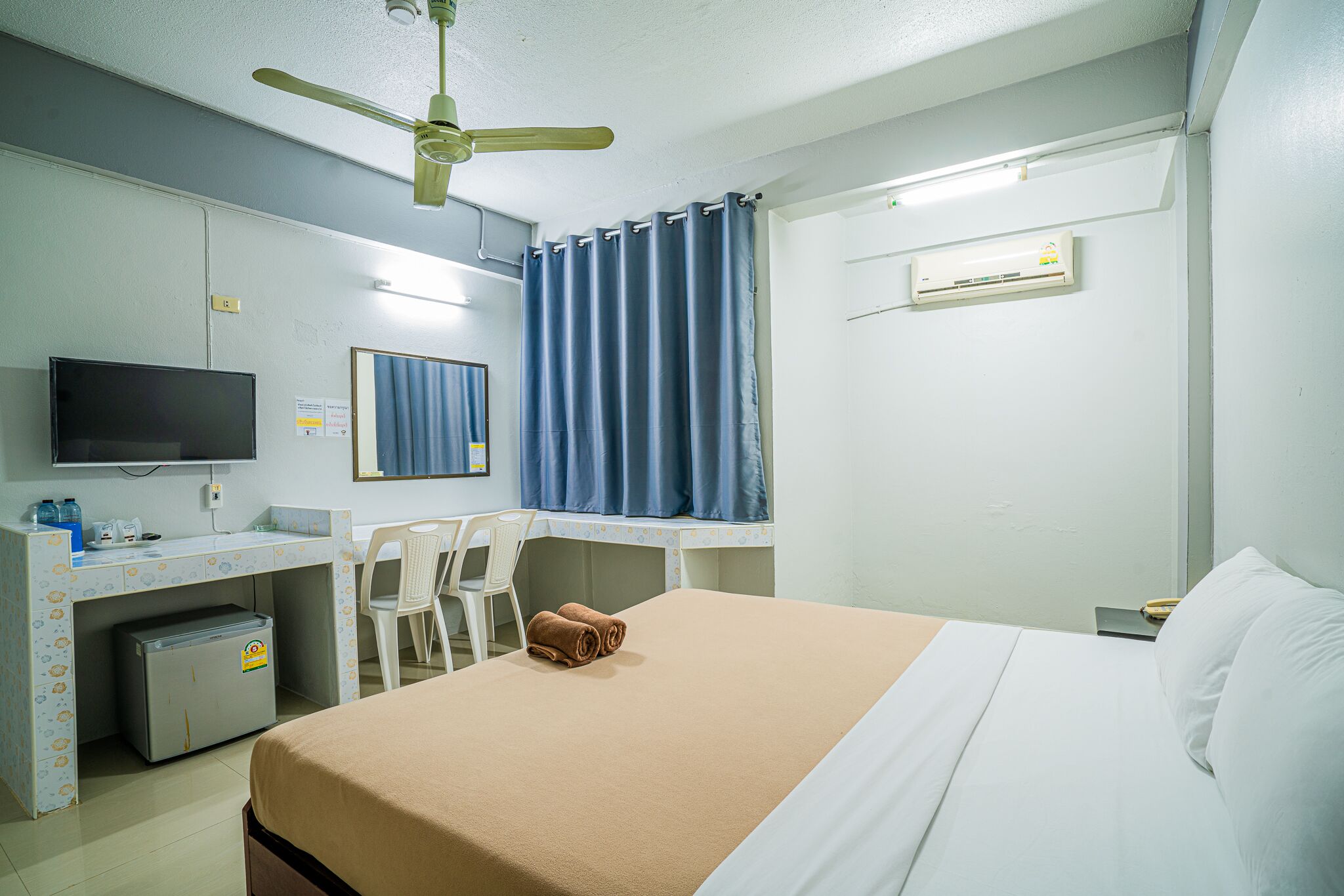 A hotel room with a bed, a desk with a chair, a television, a ceiling fan, and a window with curtains.