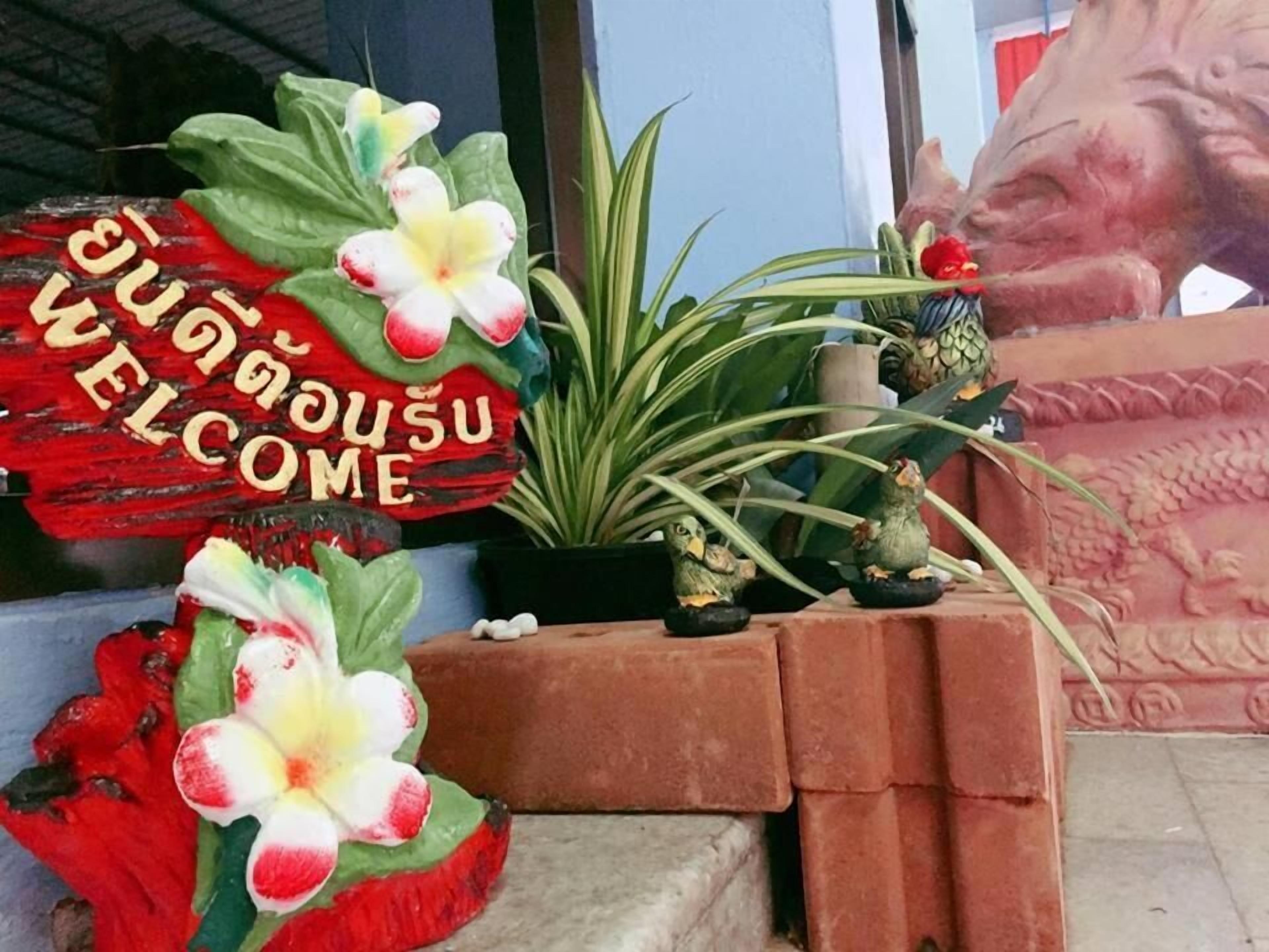 Un letrero de bienvenida con texto tailandés, una planta en maceta y aves decorativas.