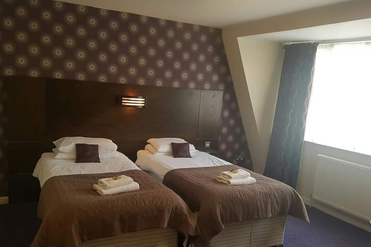 Habitación de hotel con dos camas, cada una con cubrecama marrón y ropa blanca, cabeceros con lámparas y una ventana con cortinas.