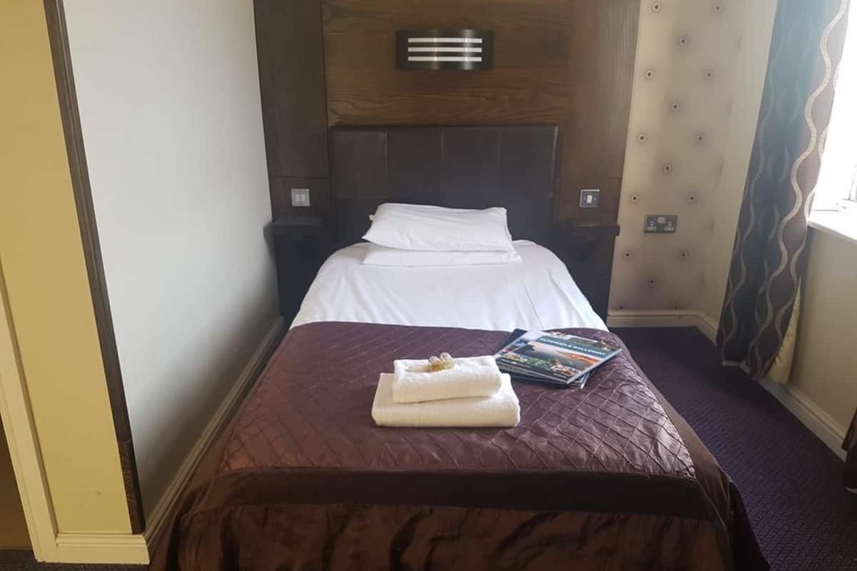 Un cuarto de hotel con una cama bien hecha, una mesita de noche con un libro y una lámpara fijada en la pared.
