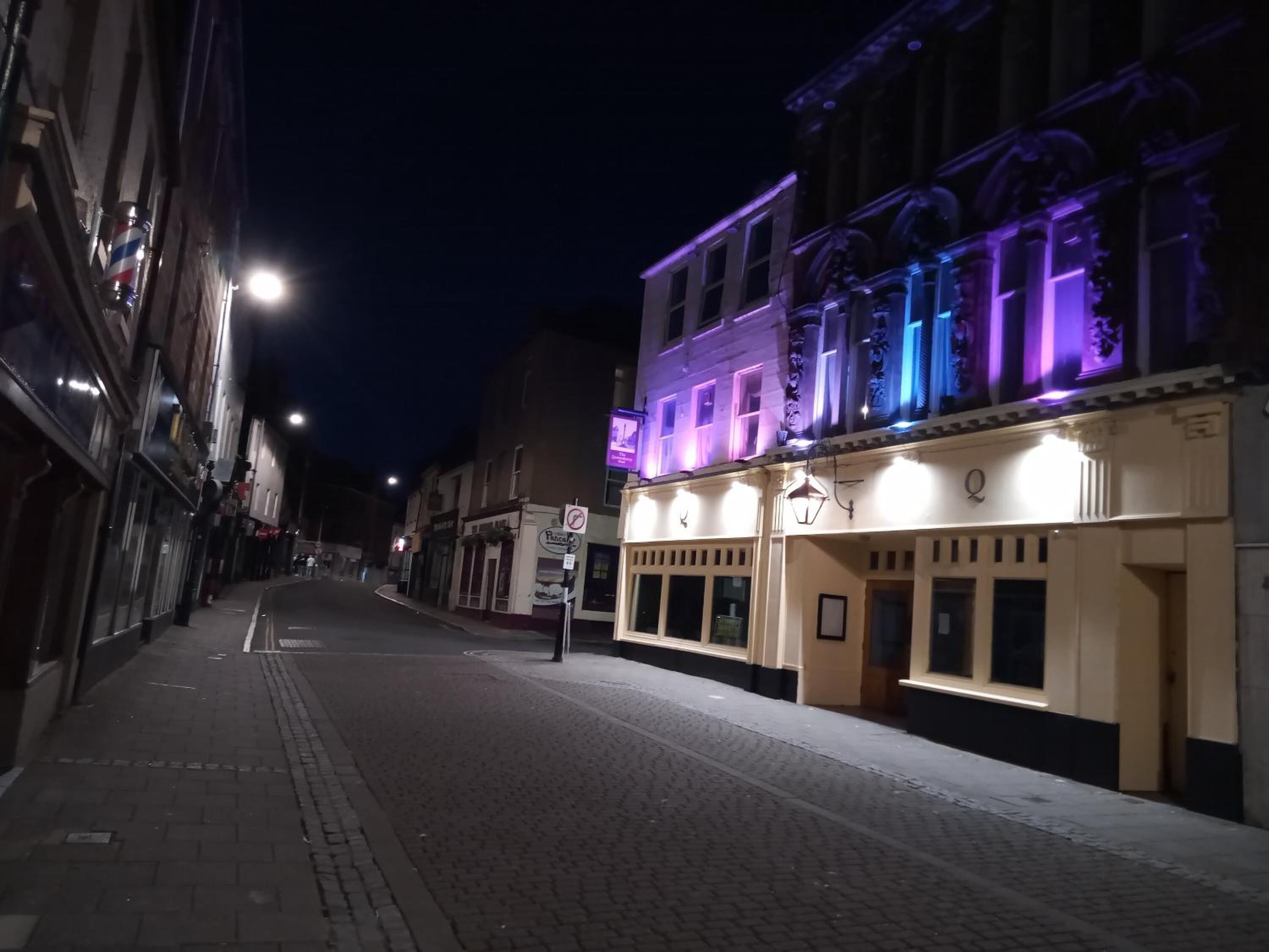 A street at night with buildings on either side, including a brightly lit building with a sign that appears to be a restaurant or cafe.