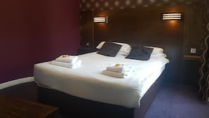 A neatly made bed with white linens and two pillows, a wooden headboard with wall-mounted lights, and a purple carpeted floor.