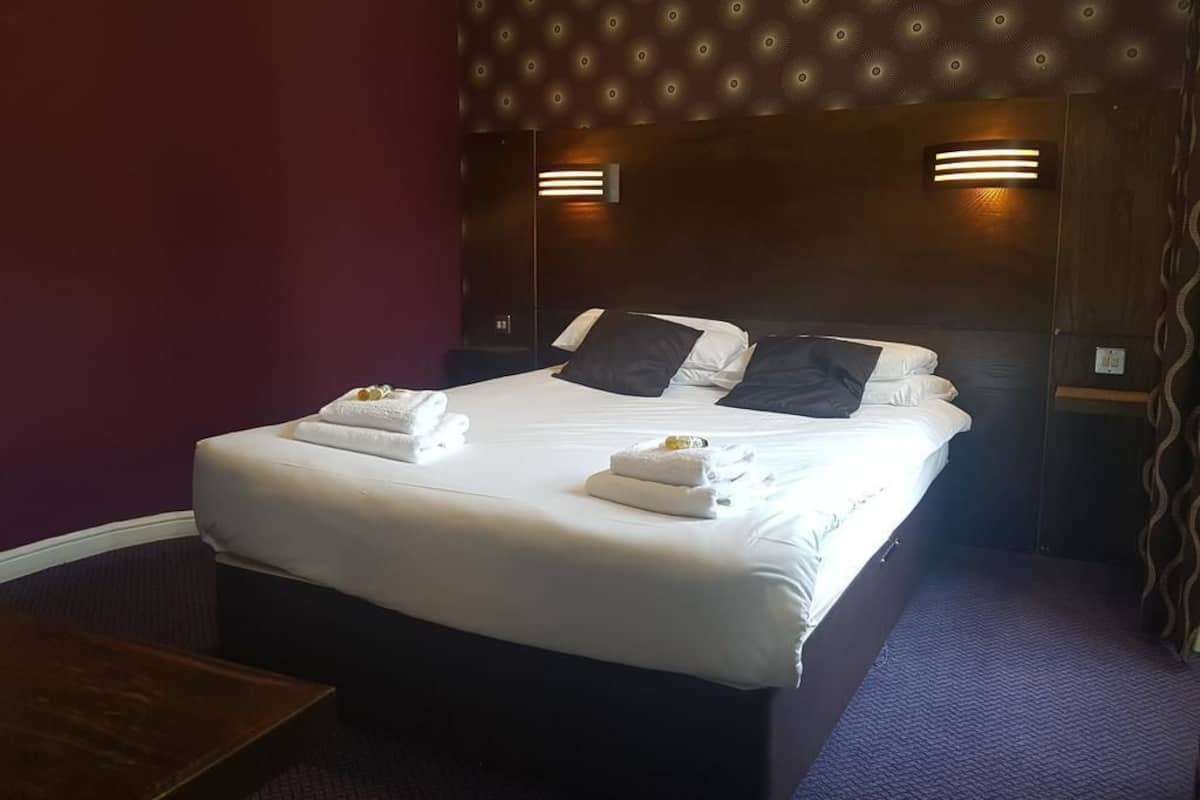 Una cama bien hecha con sábanas blancas y dos almohadas, un cabecero de madera con luces fijadas en la pared y un suelo cubierto de moqueta morada.