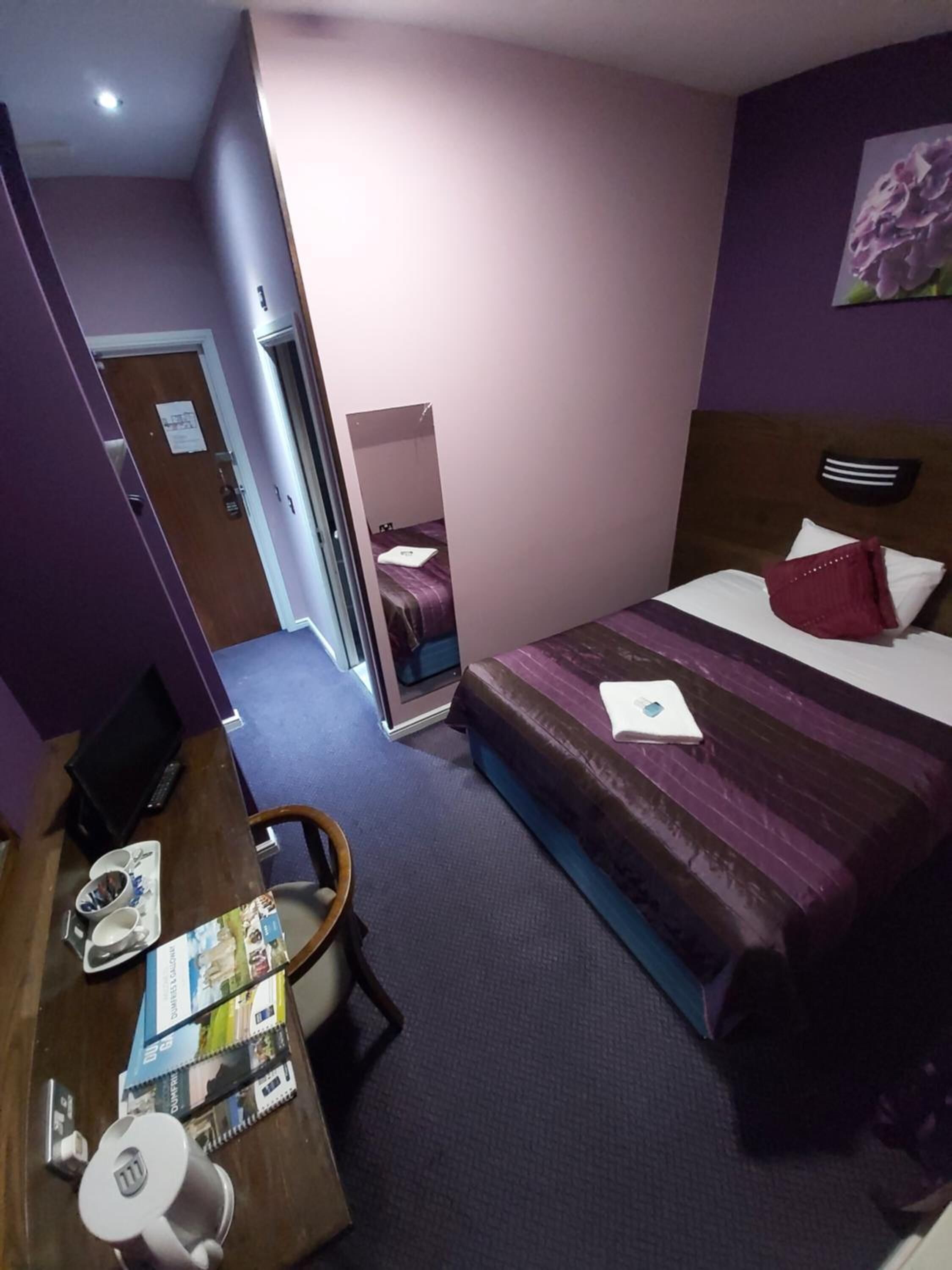 A hotel room with a bed, a desk, a chair, a mirror, and a door.
