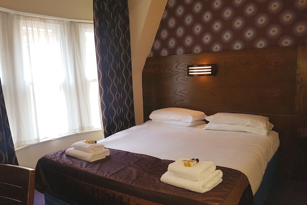 Habitación de hotel con una cama grande, cabecera con diseño estampado, ventana con cortinas translúcidas y mesita de noche con una botella y una toalla.