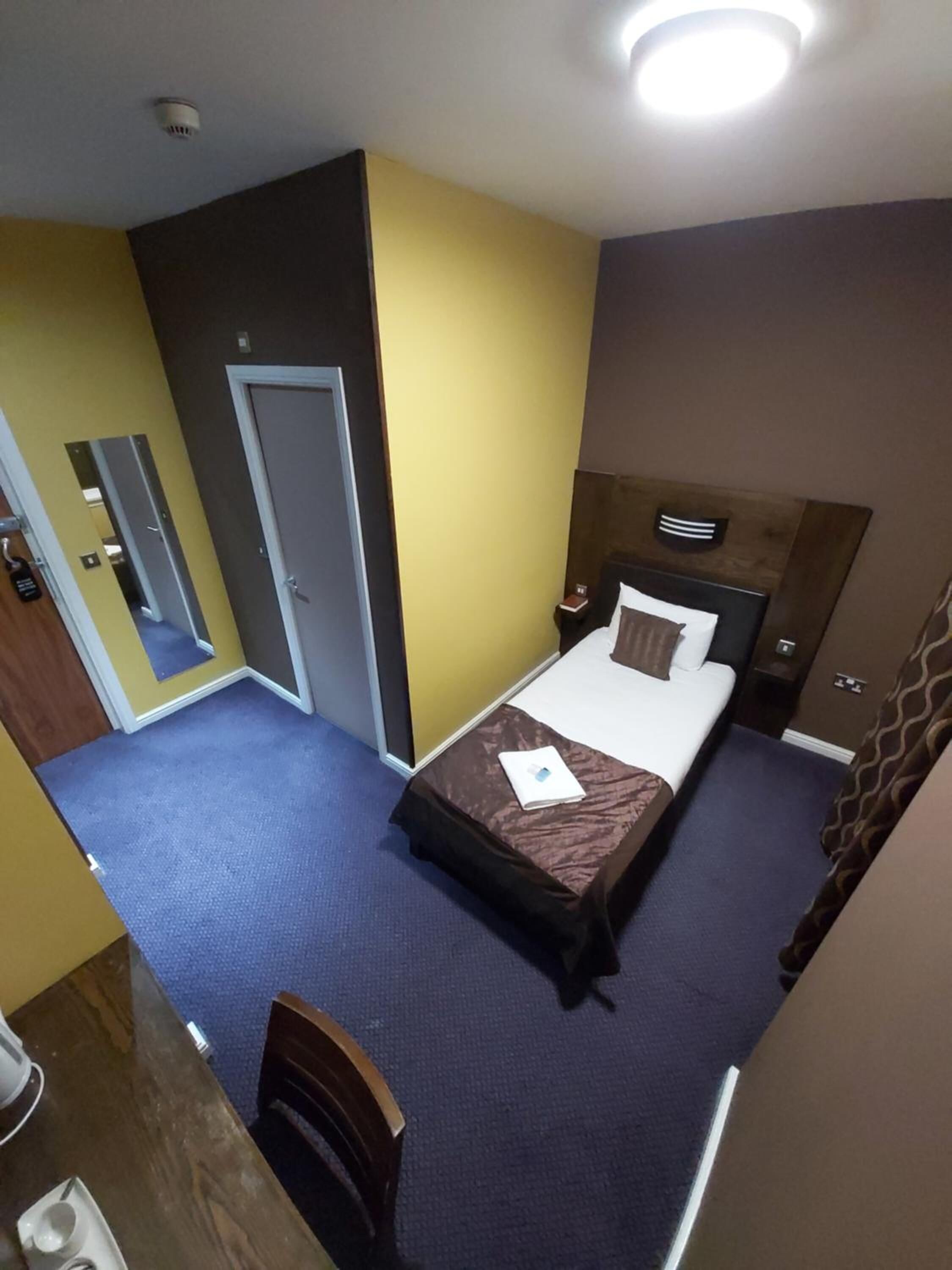 A hotel room with a single bed, a desk with a chair, a mirror, and a door.