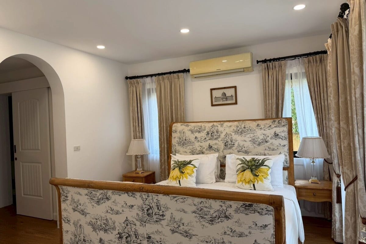 A bedroom with a bed, two bedside tables, a nightstand, a wall-mounted air conditioner, and a framed picture on the wall.