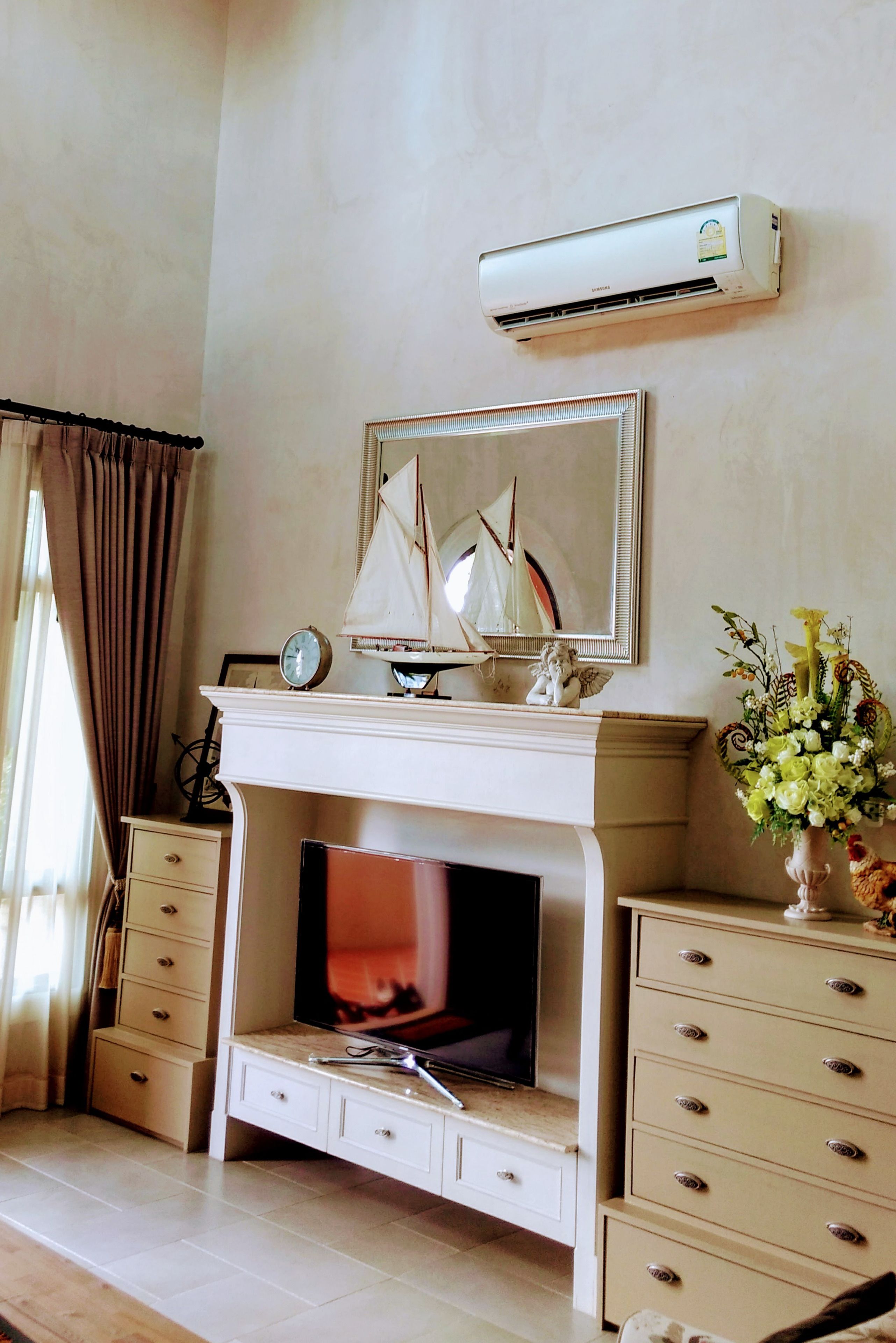 A living room with a fireplace, a mounted air conditioner, a decorative sailboat model, and a vase of flowers.