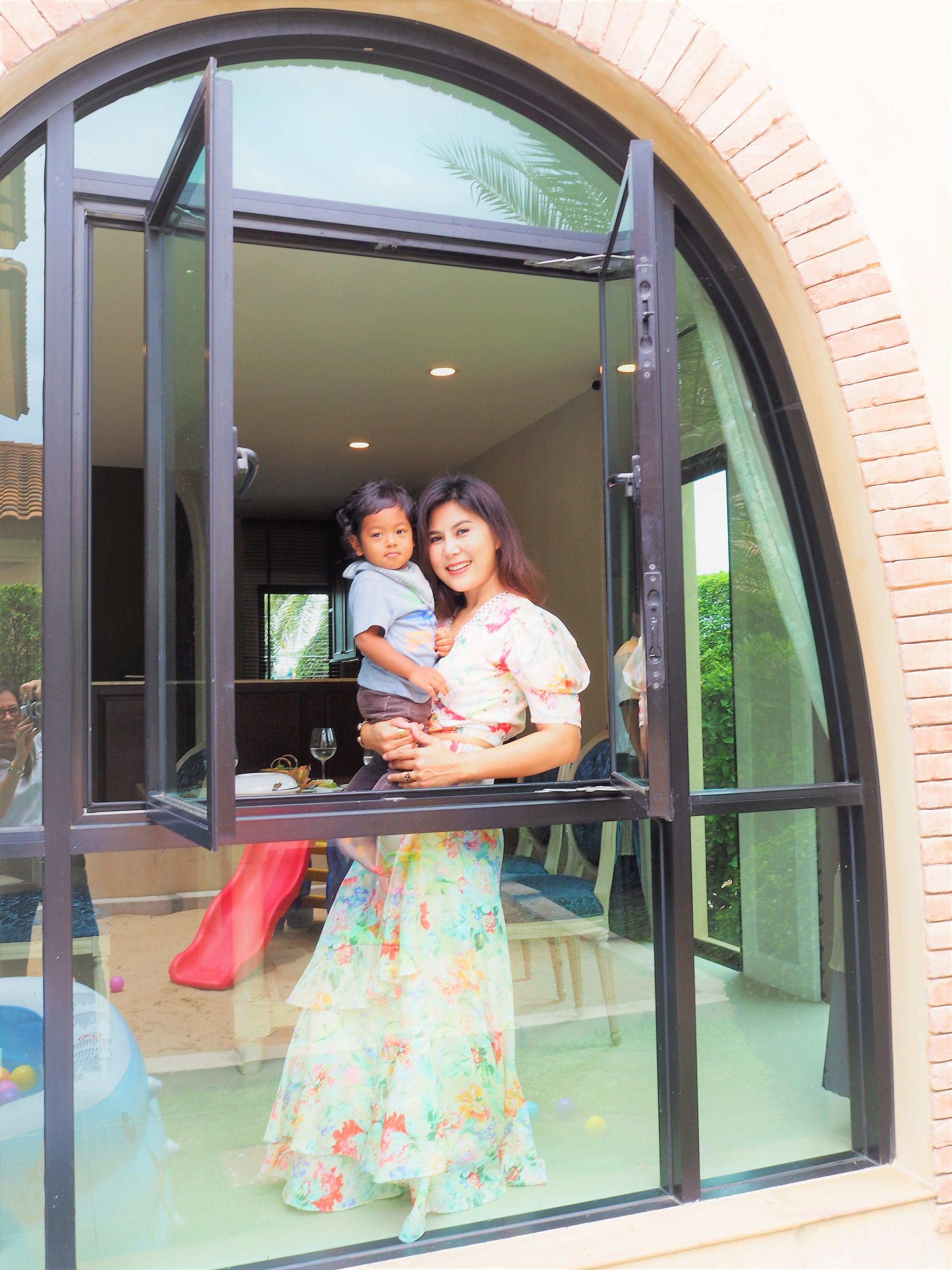 A woman holding a child in a room with a large arched window, a slide, and a pool.
