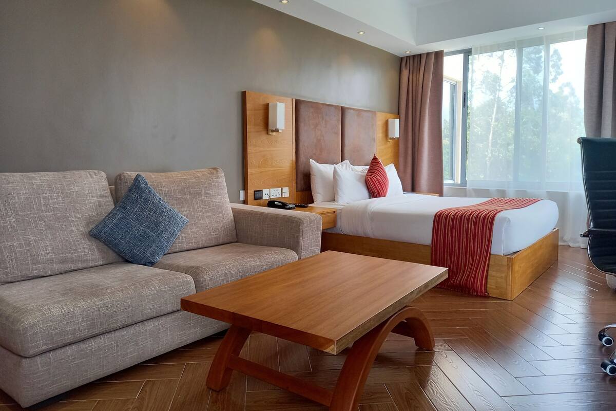 Une chambre d’hôtel avec un canapé, une table basse en bois, un lit recouvert d’une couverture rouge et une table de chevet avec un téléphone et une télécommande.