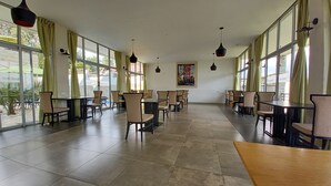 A spacious indoor area with multiple tables and chairs arranged in rows, large windows on one side, and hanging pendant lights.