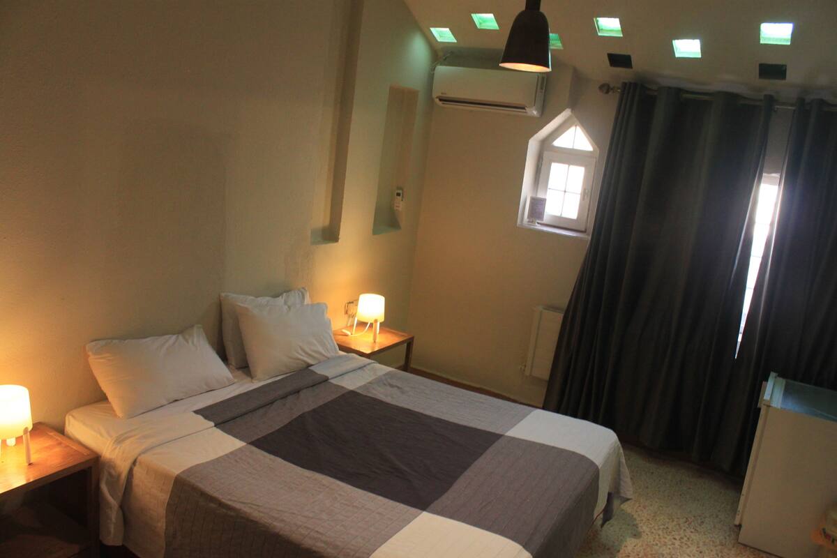 A bedroom with a bed, bedside lamps, a window with curtains, and an air conditioning unit.