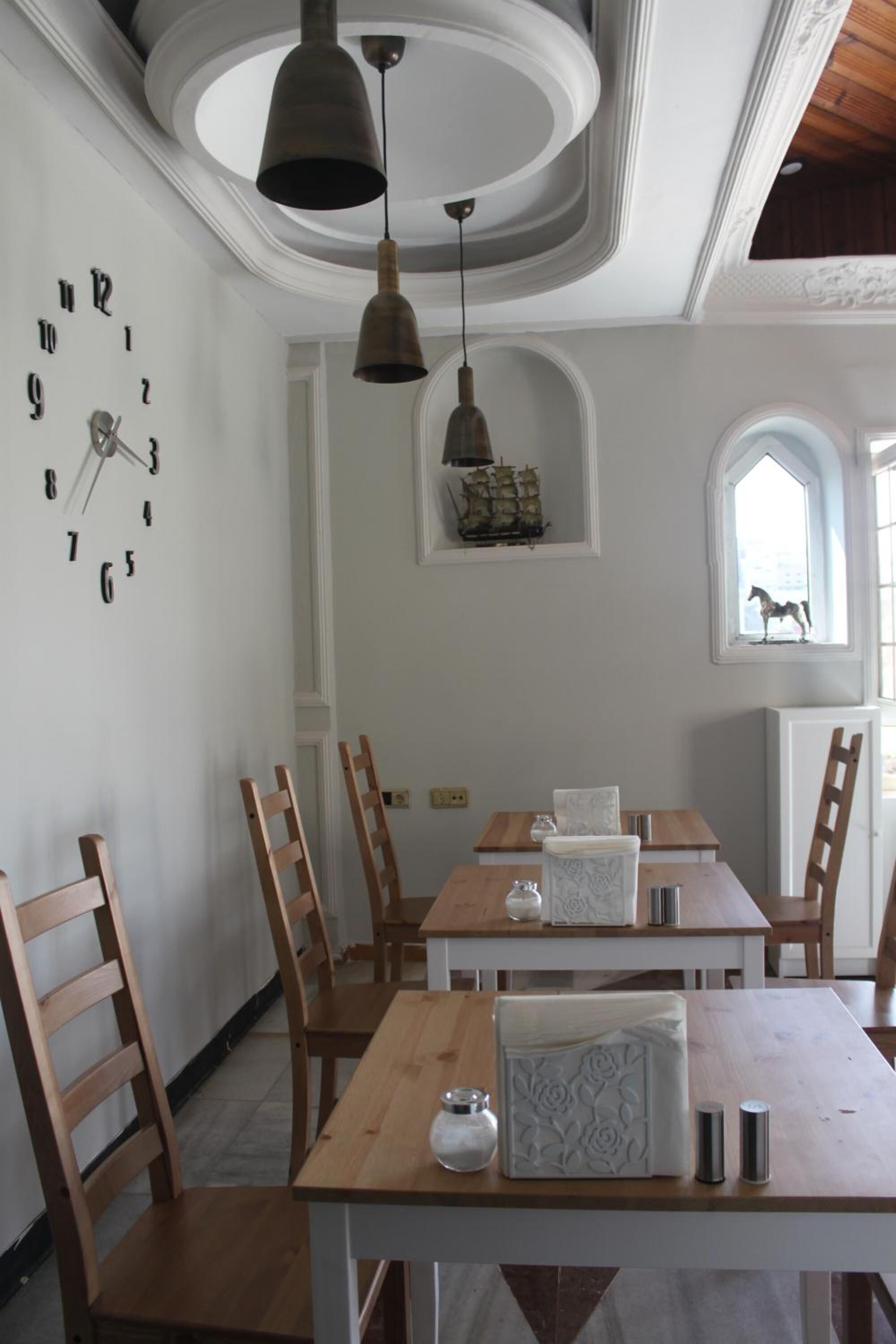 Un comedor con mesas y sillas de madera, un reloj de pared, luces colgantes y una ventana con vistas.