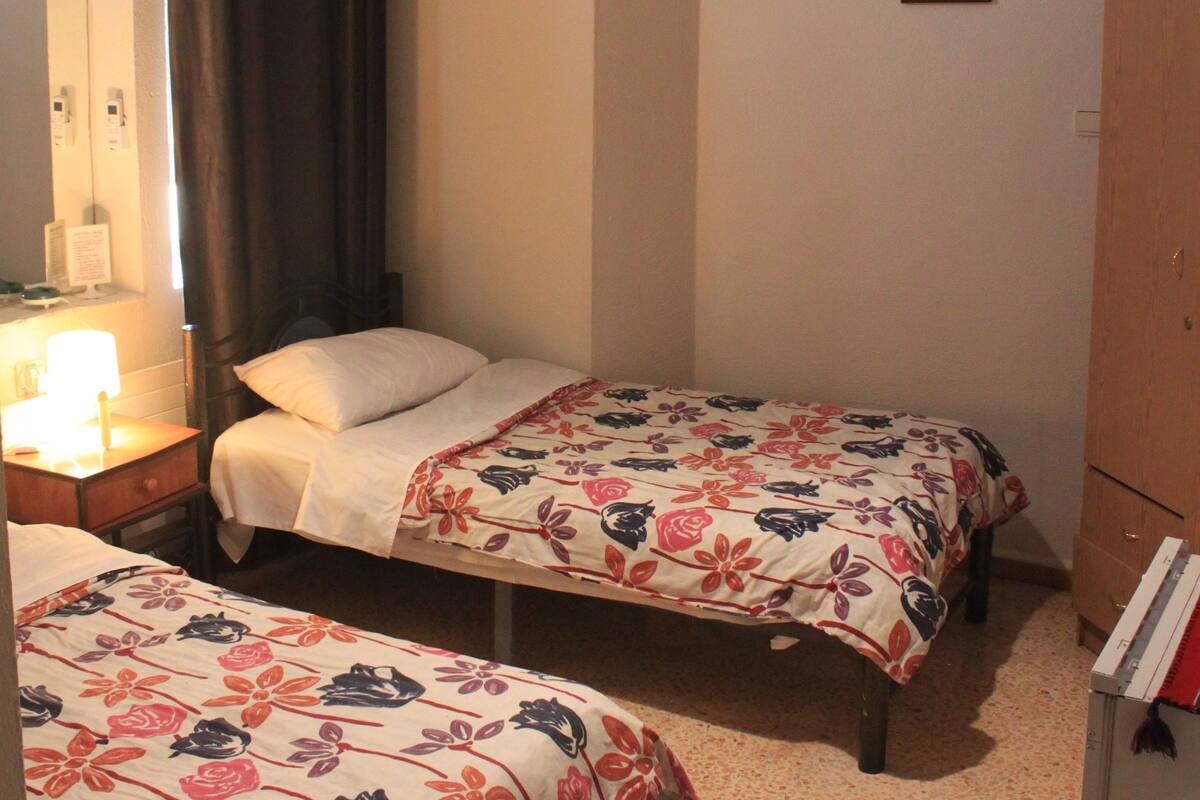 A hotel room with two single beds, a bedside table with a lamp, a wardrobe, and a small desk with a chair.