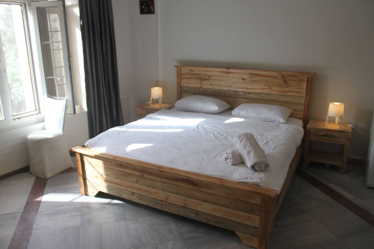 A wooden bed with white linens, two bedside tables with lamps, and a window with curtains.