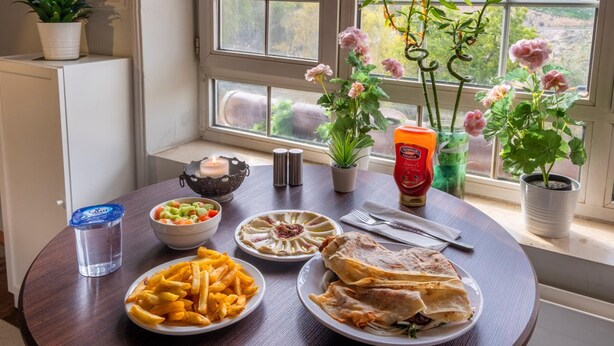 A dining table with plates of food, a candle, and potted plants by a window with a city view. - Cairwan Hotel (Al-Karak)