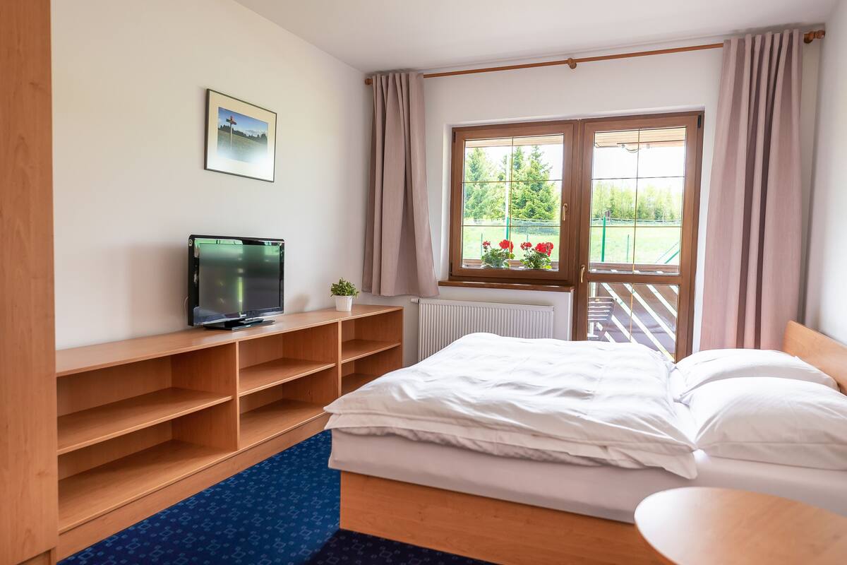 Un dormitorio con cama, televisor sobre una estantería de madera, ventana con cortinas y radiador.