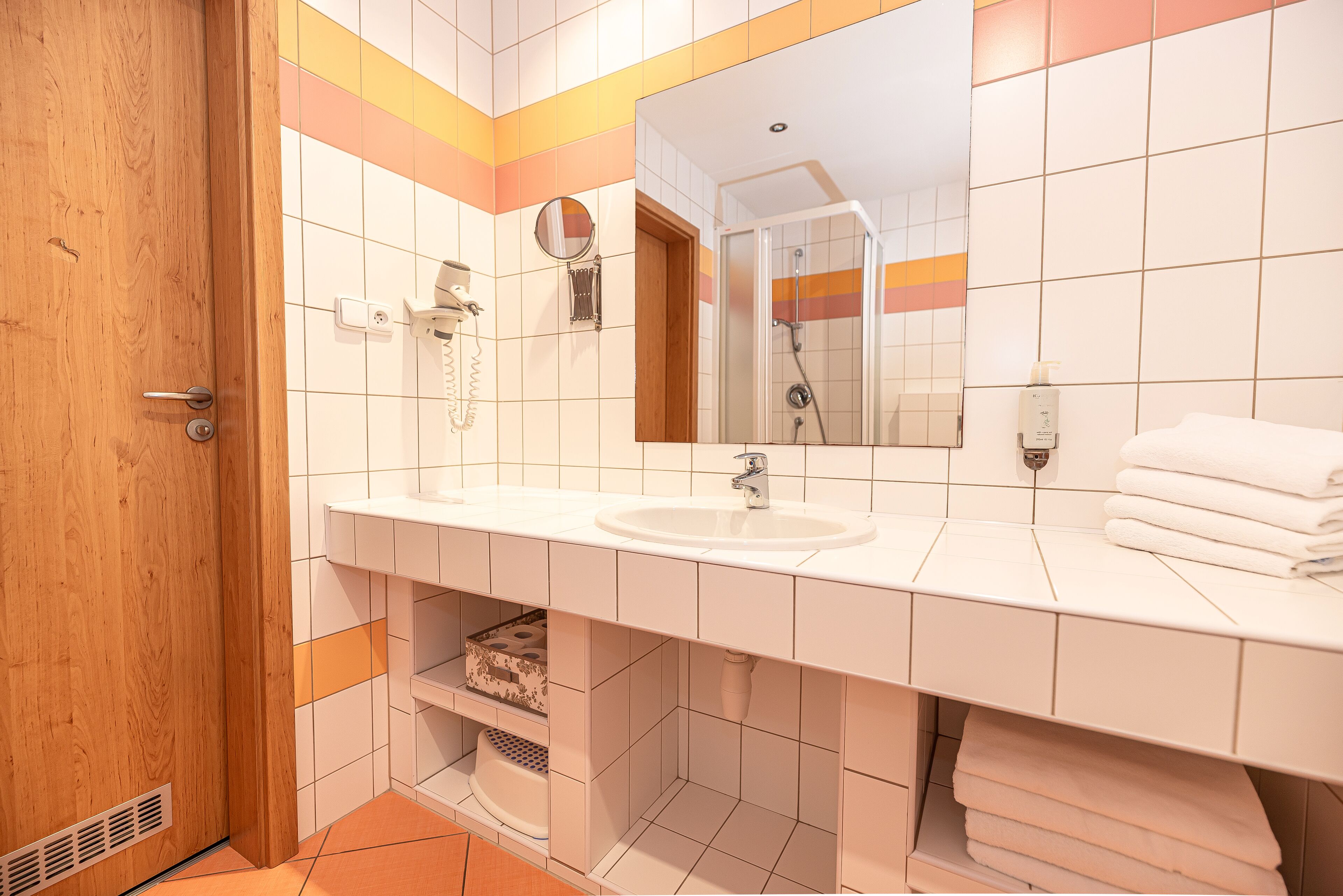 Un baño con puerta de madera, azulejos blancos, un lavabo, un espejo y un perchero para toallas.