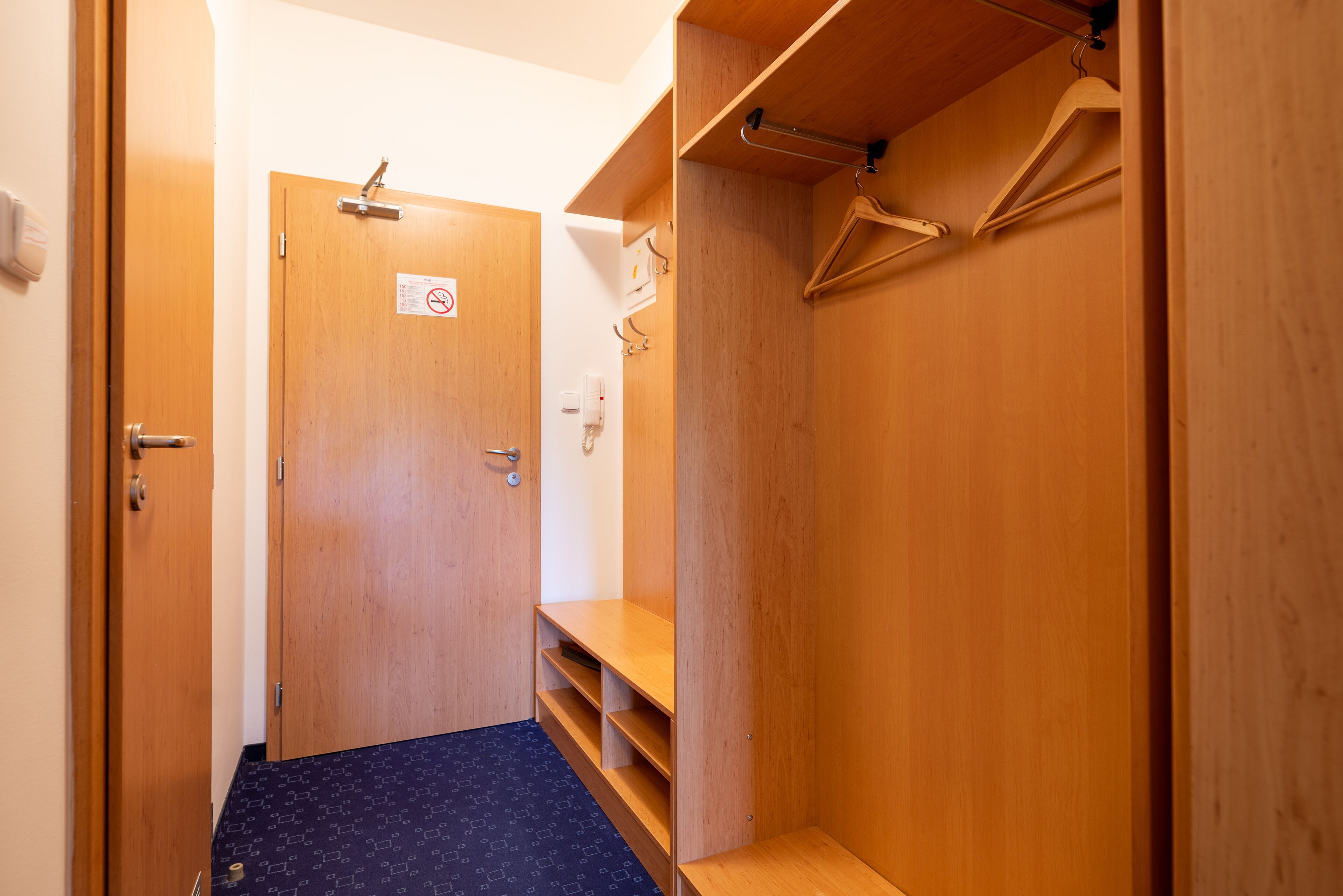 A wooden closet with shelves and a hanging rod, a door with a 'no smoking' sign, and a blue patterned carpet.