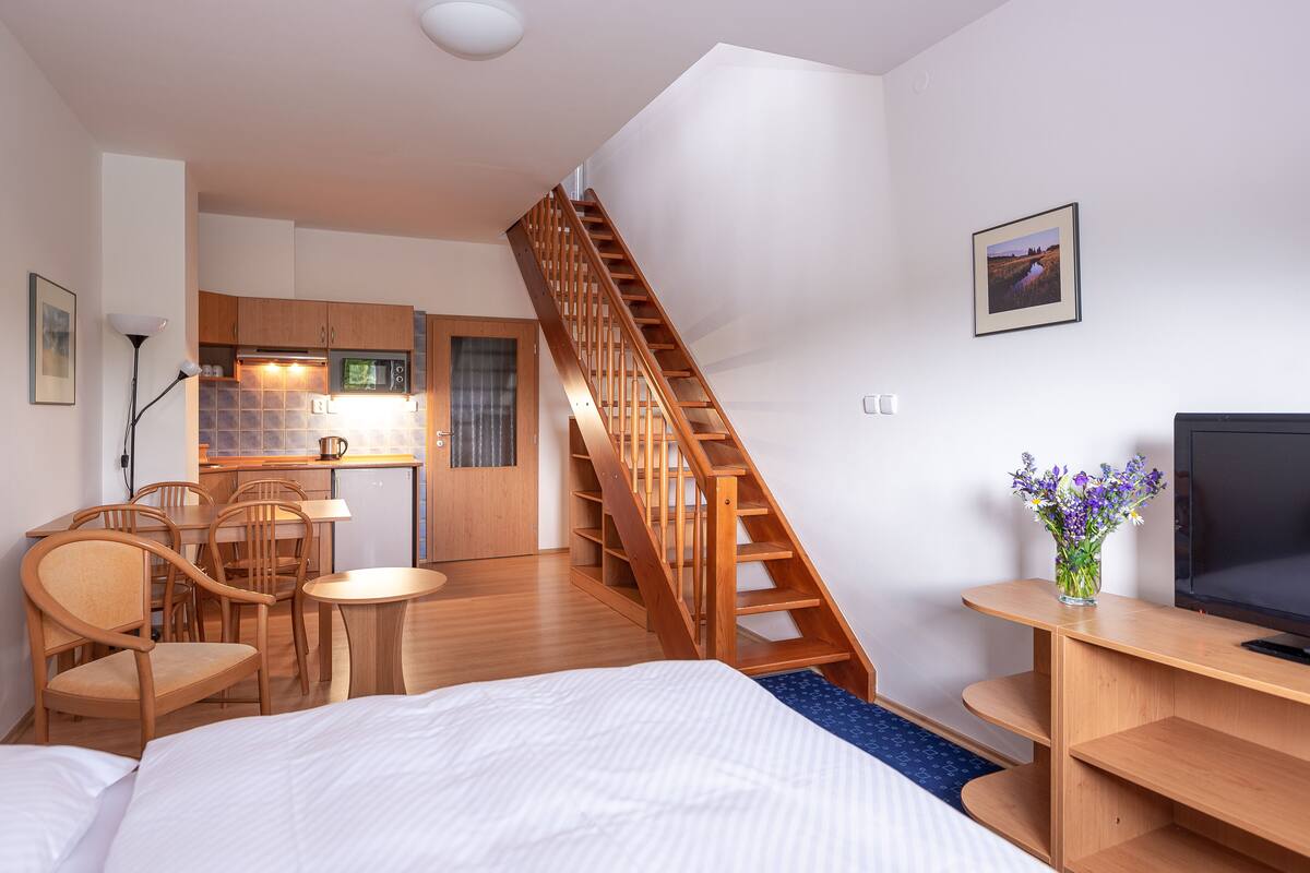 Una habitación de hotel compacta con una cama, un escritorio, una silla, una mesita, un televisor, una escalera de madera y un rincón americano.