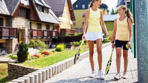 Two people walking on a paved pathway with tennis rackets, surrounded by houses with distinct architectural styles. - Apartmany Engadin (Bozi Dar)