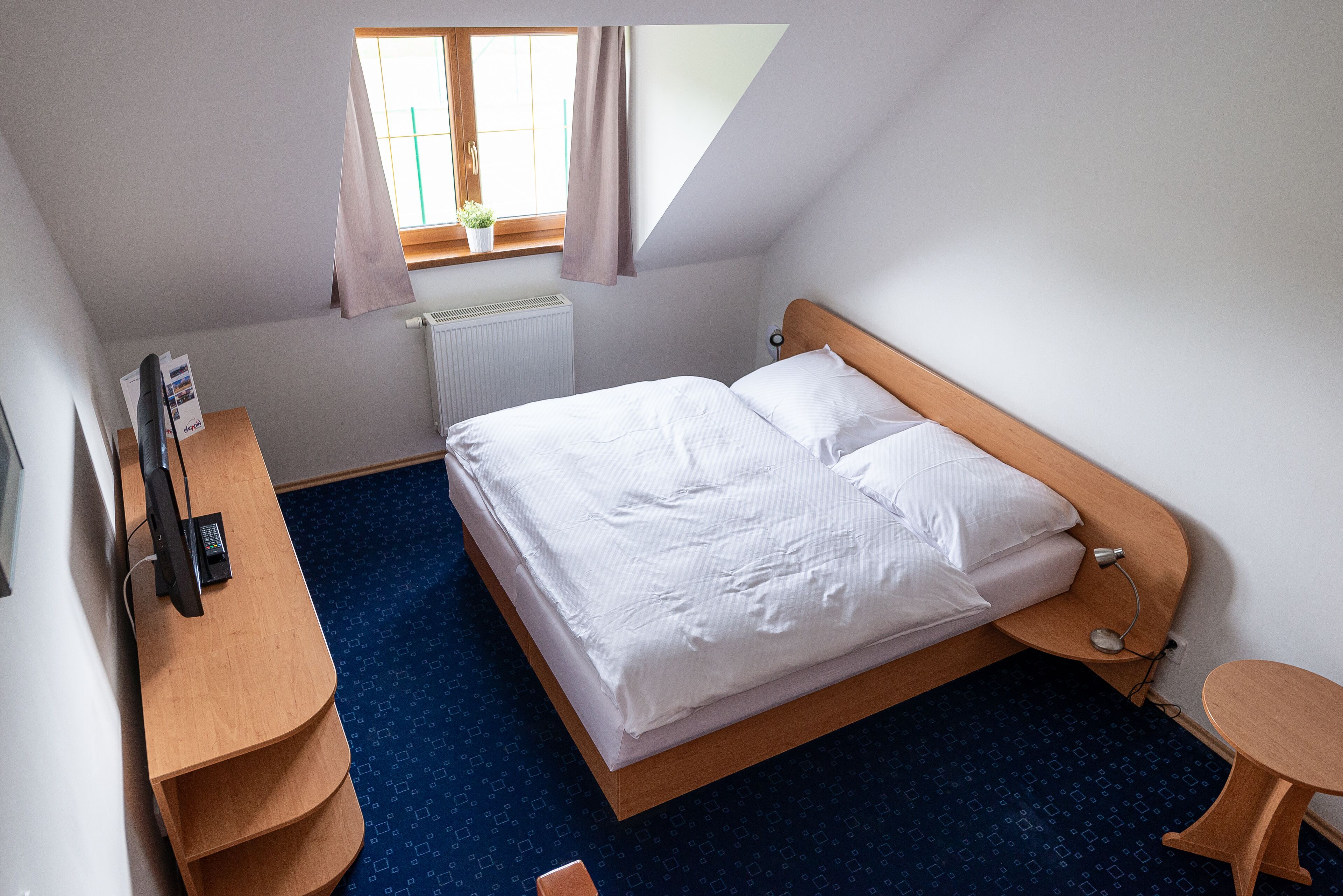A bedroom with a bed, a desk, a chair, a small table, a radiator, and a window with curtains.