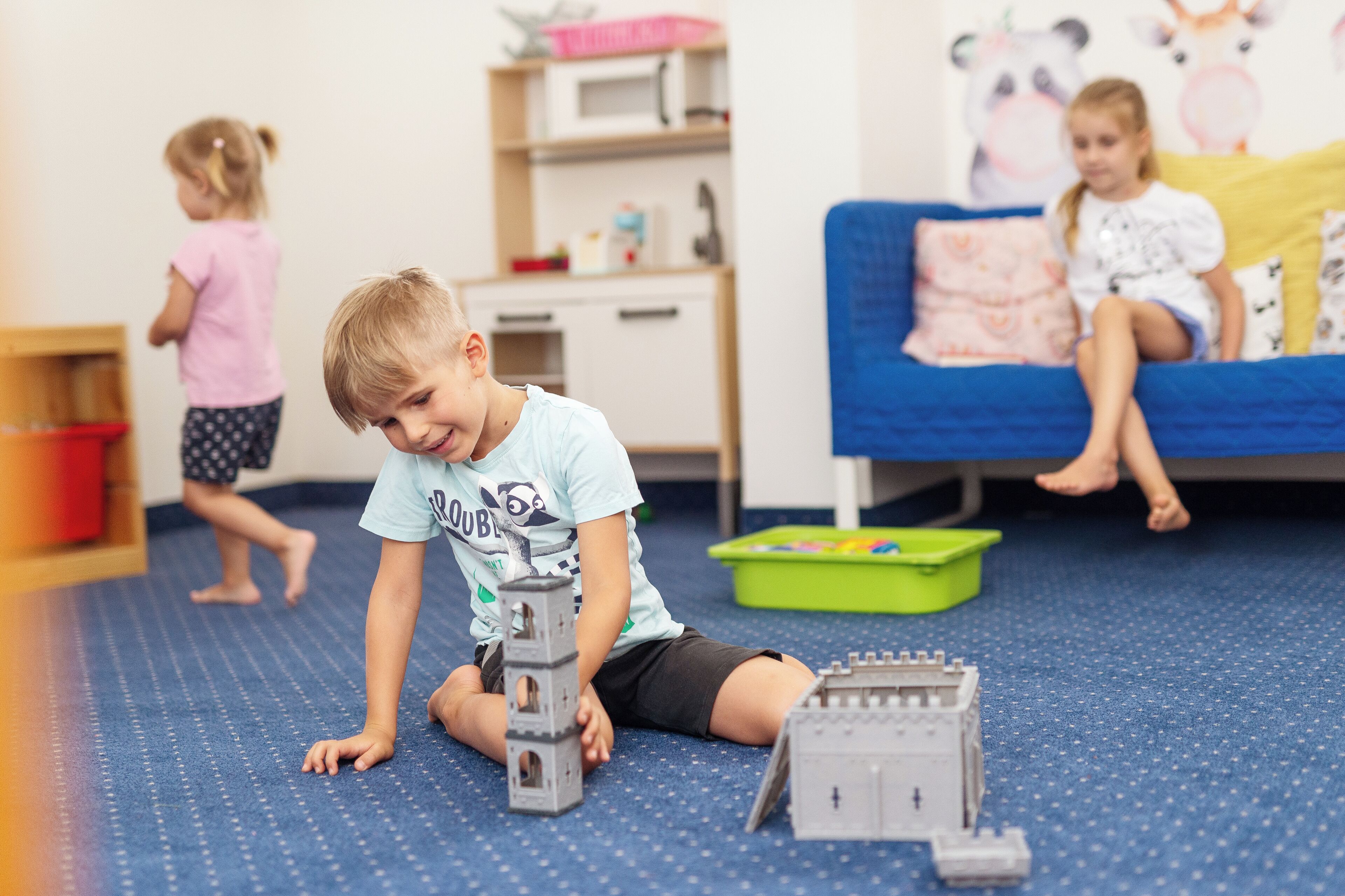 A child playing with toy structures on a blue carpet in a room with a bed and a shelf in the background.