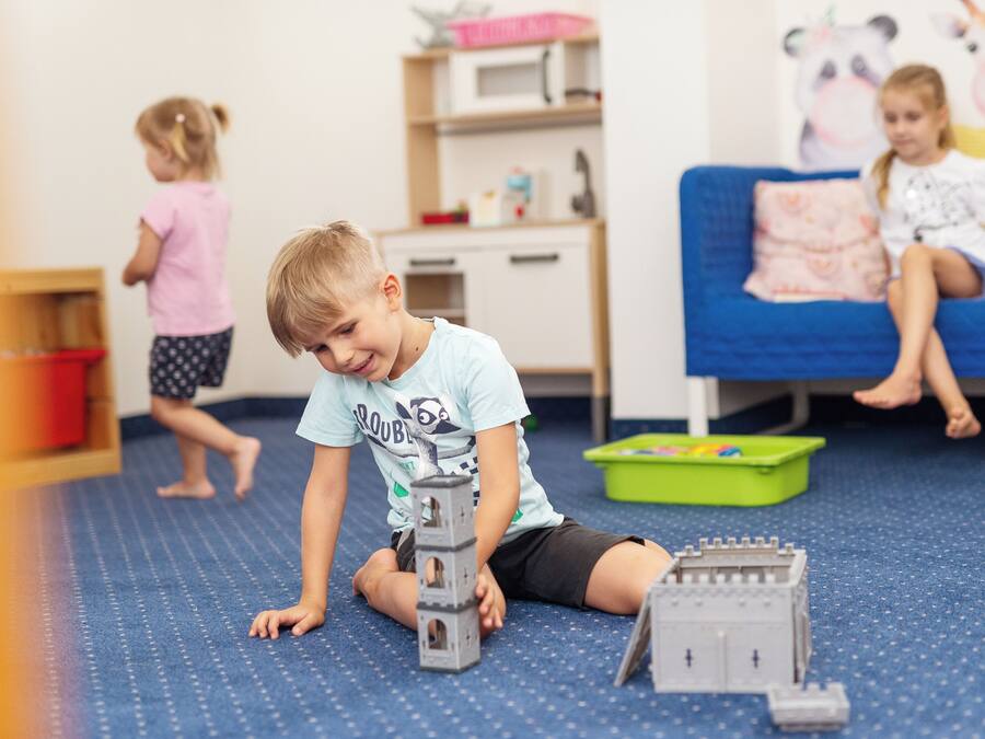 A child playing with toy structures on a blue carpet in a room with a bed and a shelf in the background.