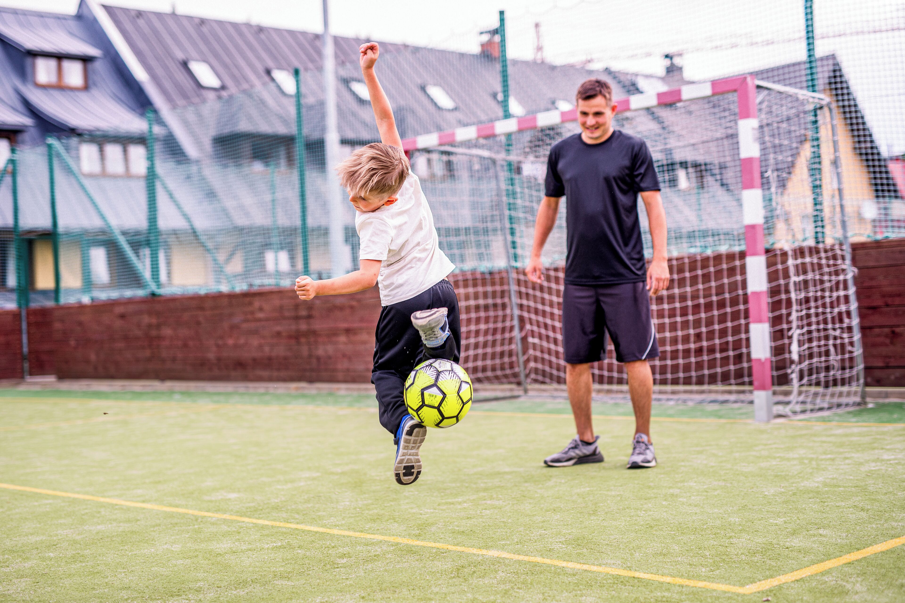 Un campo de fútbol con un arco y una red, un niño pateando un balón y un hombre parado cerca.