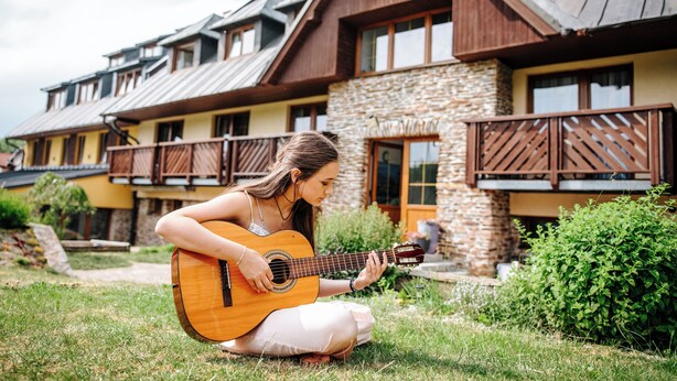 A person playing an acoustic guitar in front of a building with a stone facade and wooden balconies. - Apartmany Engadin (Bozi Dar)