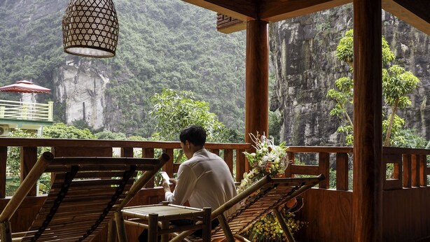 A person sitting on a wooden chair with a bamboo table, overlooking a mountainous landscape. - Trang An Heritage Garden (Tay Hoa Lu)
