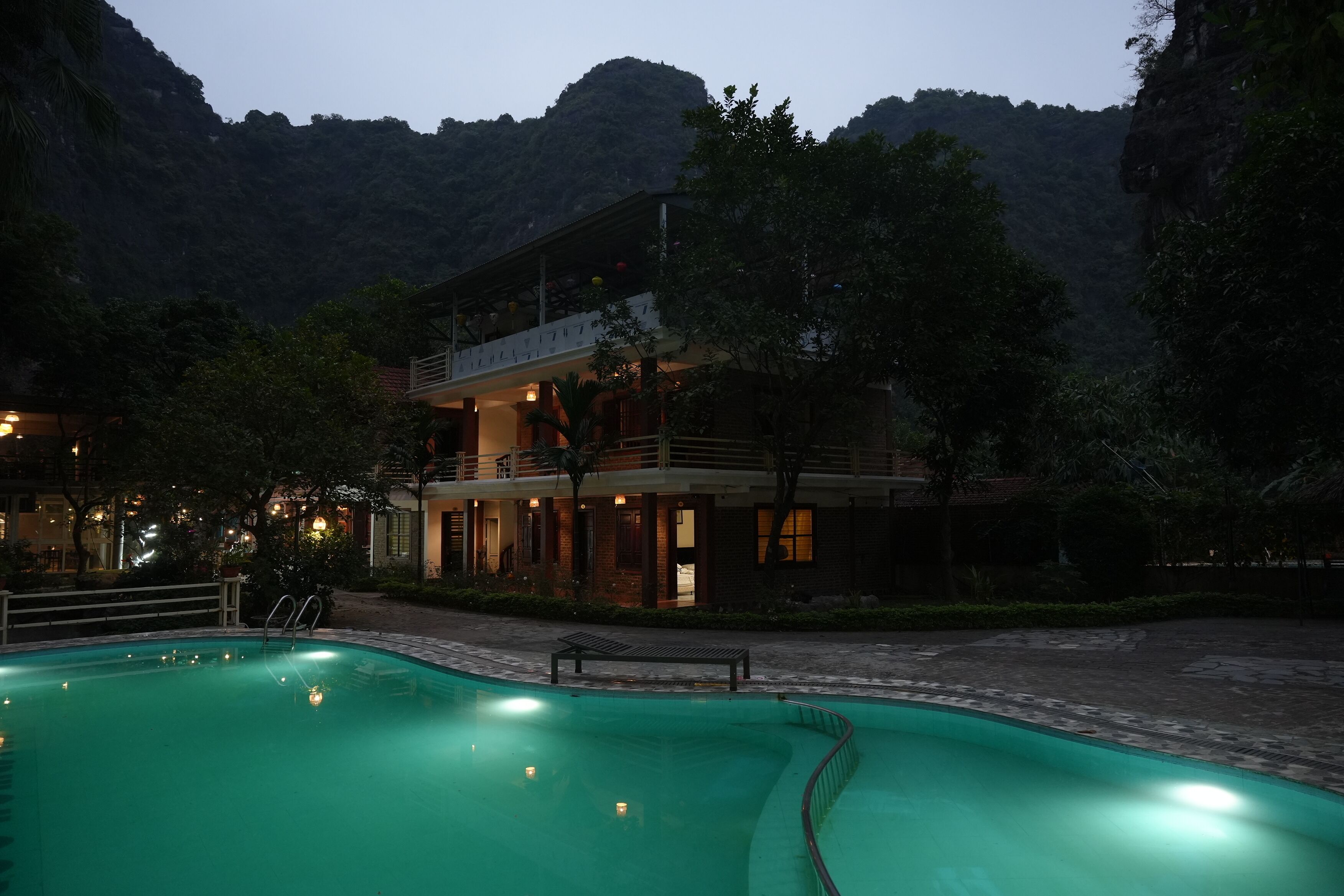A swimming pool with a curved edge, a wooden deck, and a building with a balcony in a mountainous area.