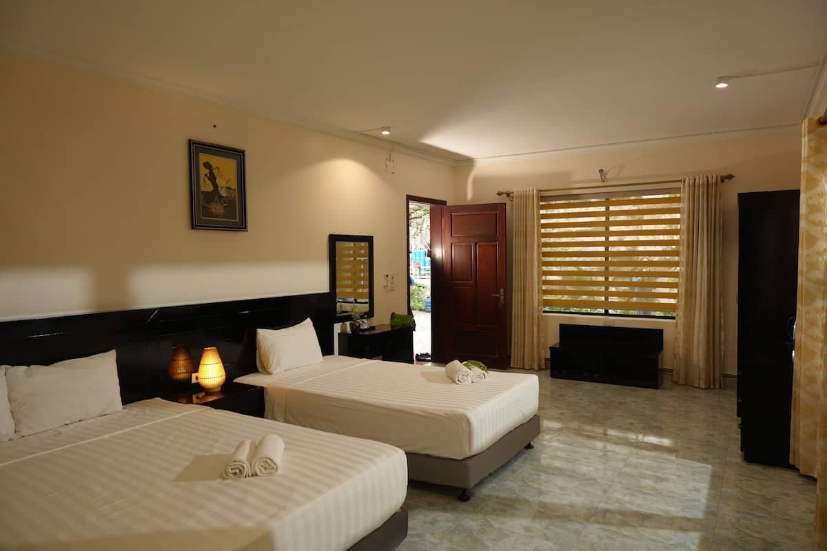 Habitación de hotel con dos camas, una mesita de noche con lámpara, una puerta de madera y una ventana con persianas.