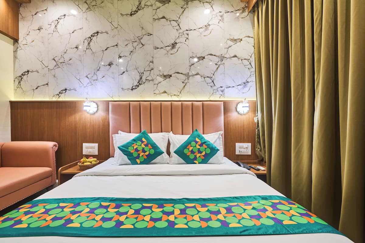 A hotel room with a marble-patterned wall, a bed with white bedding, two pillows with a colorful pattern, a wooden headboard, and a brown sofa.