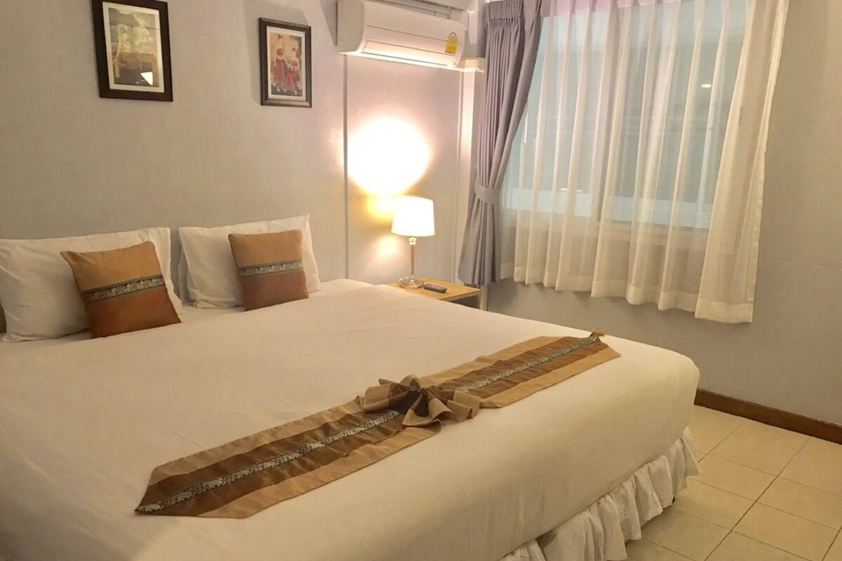 A neatly made bed with a decorative runner, a bedside lamp, and a window with sheer curtains.