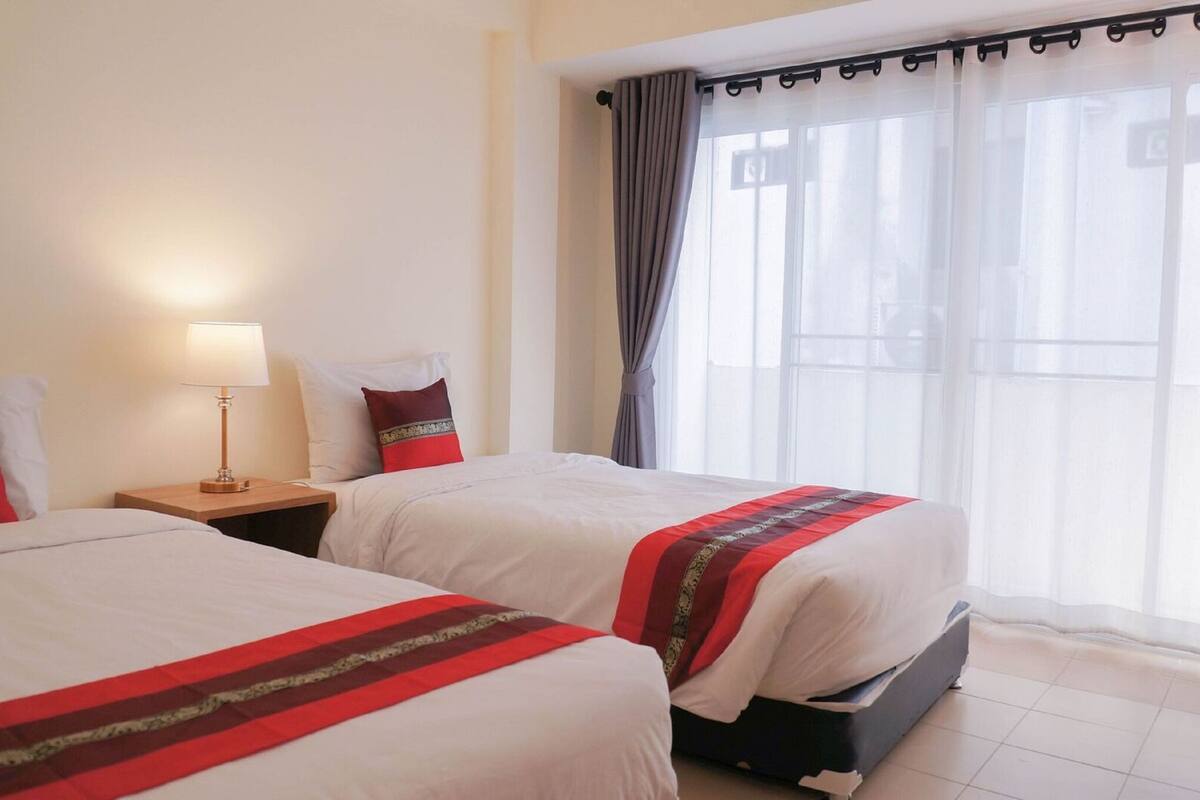 A hotel room with two beds, a nightstand with a lamp, and a large window with curtains.