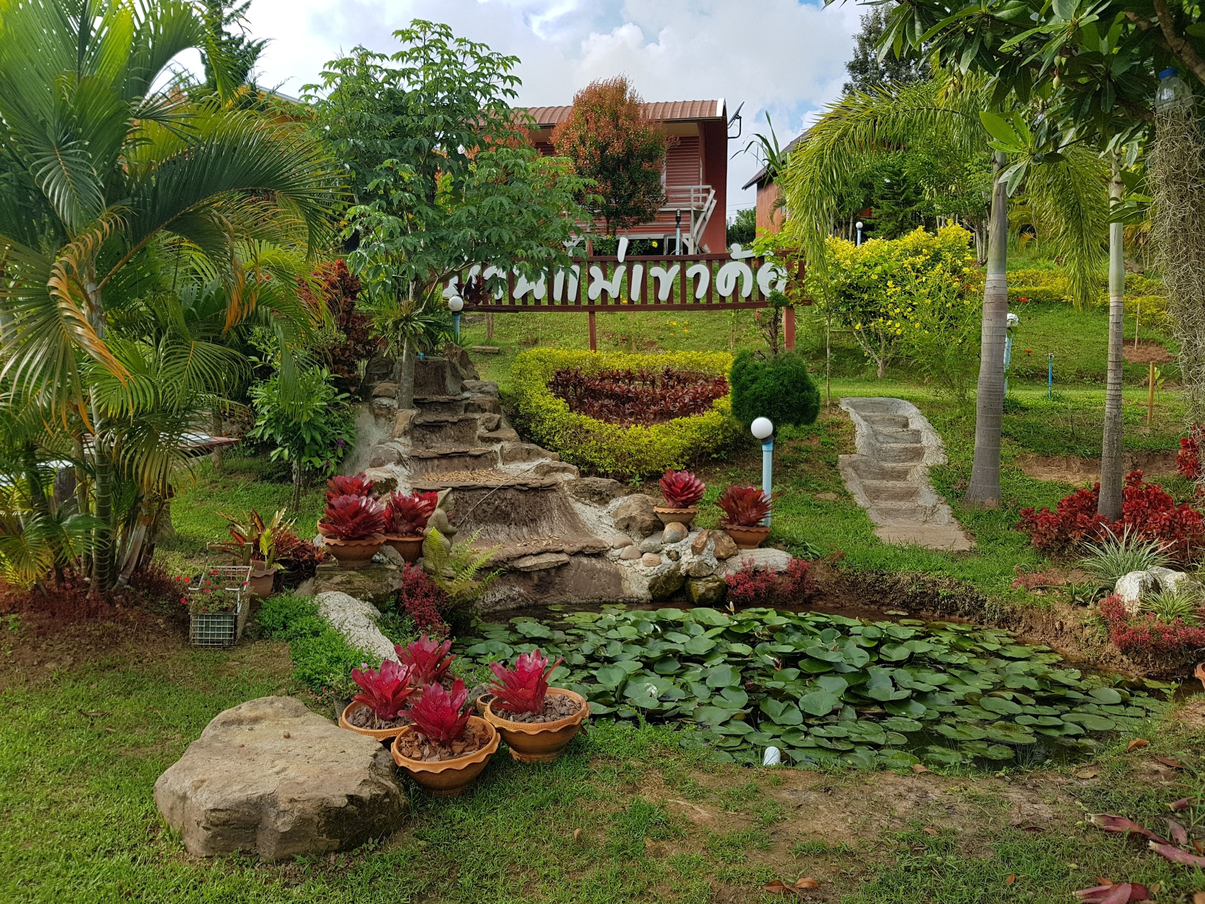 Un jardin bien entretenu, agrémenté d’une fontaine en pierre, de plantes en pot et d’un sentier menant à un bâtiment sur lequel est apposée une enseigne dans une langue étrangère.