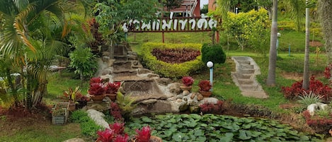 A well-maintained garden with a stone water feature, potted plants, and a pathway leading to a building with a sign in a foreign language.