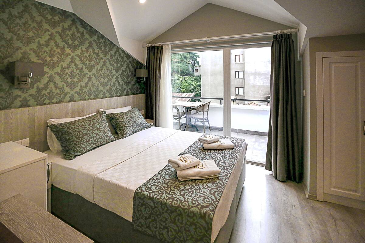 A bedroom with a large bed, a nightstand, a window with curtains, and a view of the outside balcony.