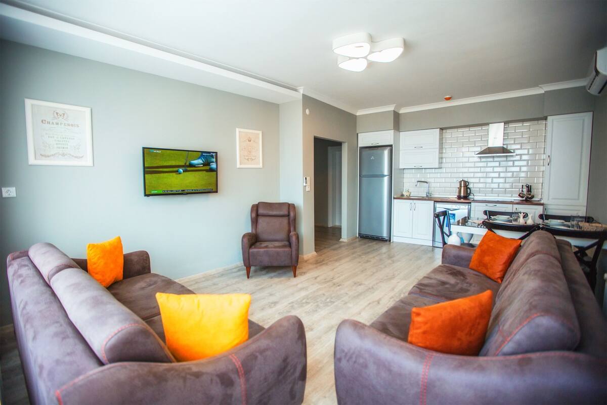 A modern living room with a sofa set, a television, and a kitchen in the background.