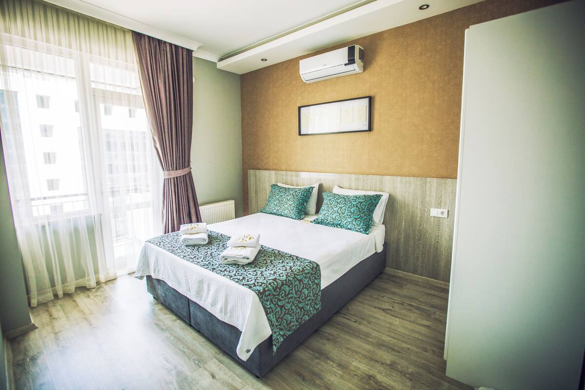 A hotel room with a bed, curtains, an air conditioning unit, and a view of buildings outside.