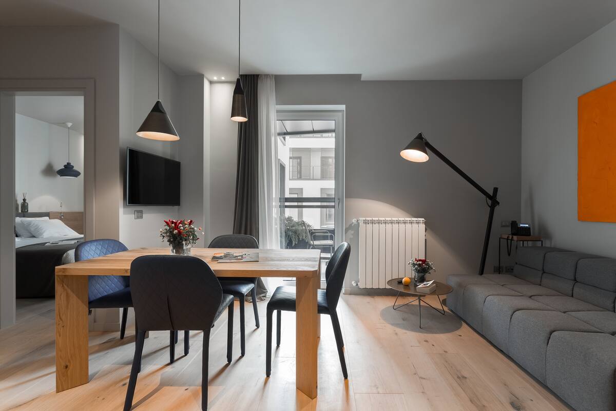 A modern living room with a wooden dining table, black chairs, a grey sofa, and a TV mounted on the wall.
